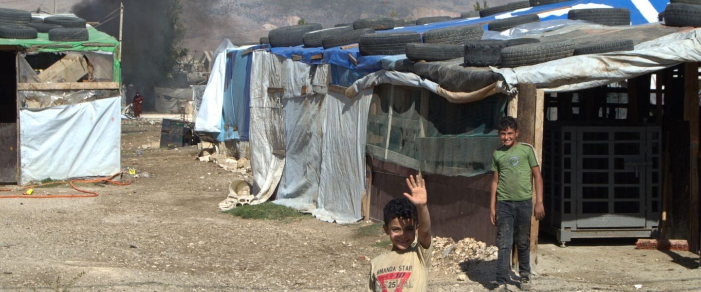 A little boy waves goodbye inside the Jurahiya settlement in Lebanon’s West Beqaa Valley as smoke from a trash fire drifts across the camp.